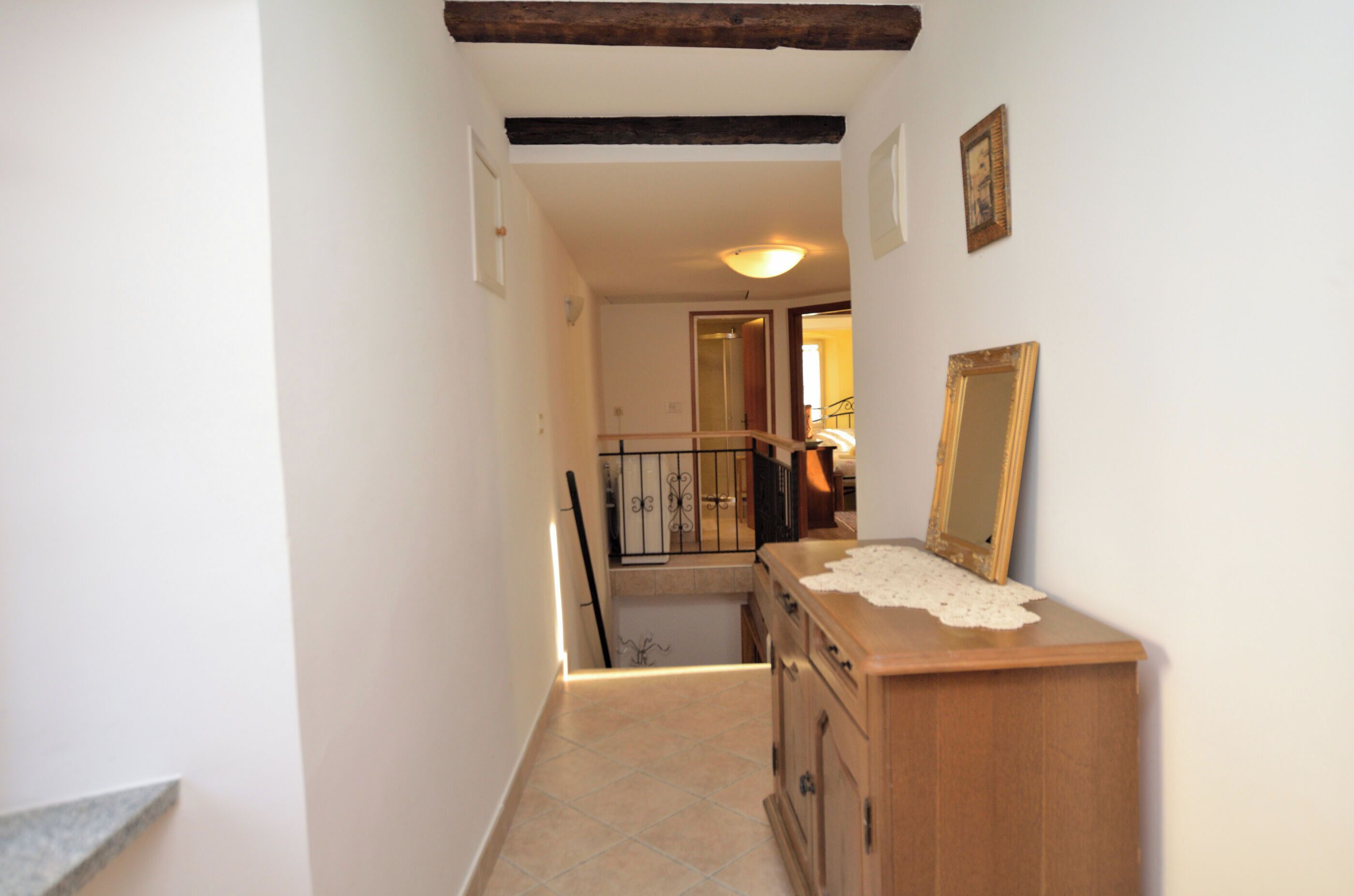 A narrow hallway with a wooden staircase, a wooden dresser with a mirror, and a tiled floor.