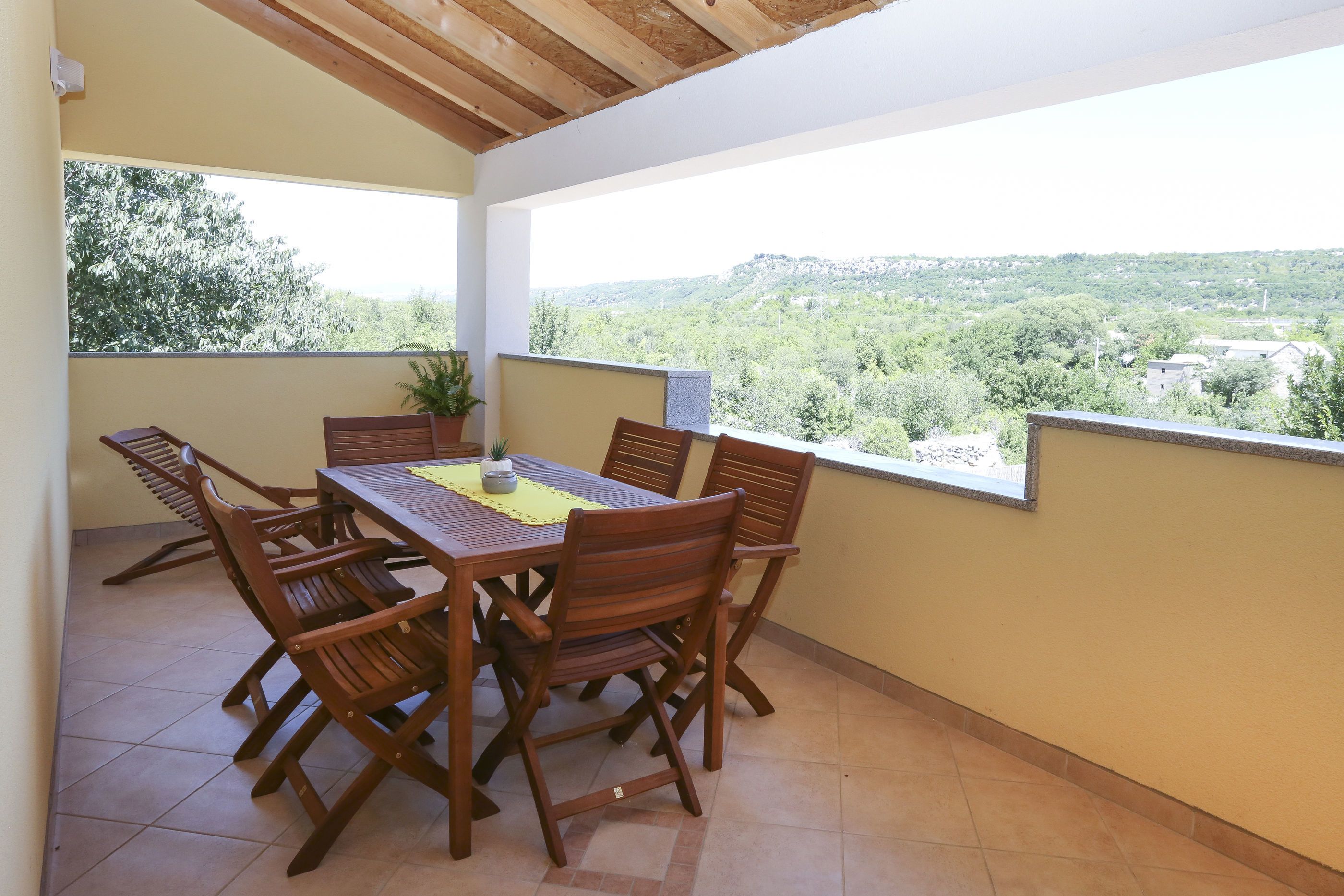 A covered patio with a wooden table and chairs, a potted plant, and a view of a hilly landscape.