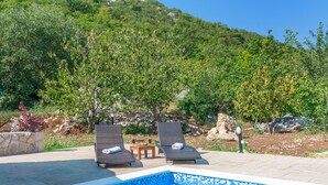 Ein Poolbereich mit zwei Liegestühlen und einem kleinen Tisch, umgeben von üppiger Vegetation und blauem Himmel.