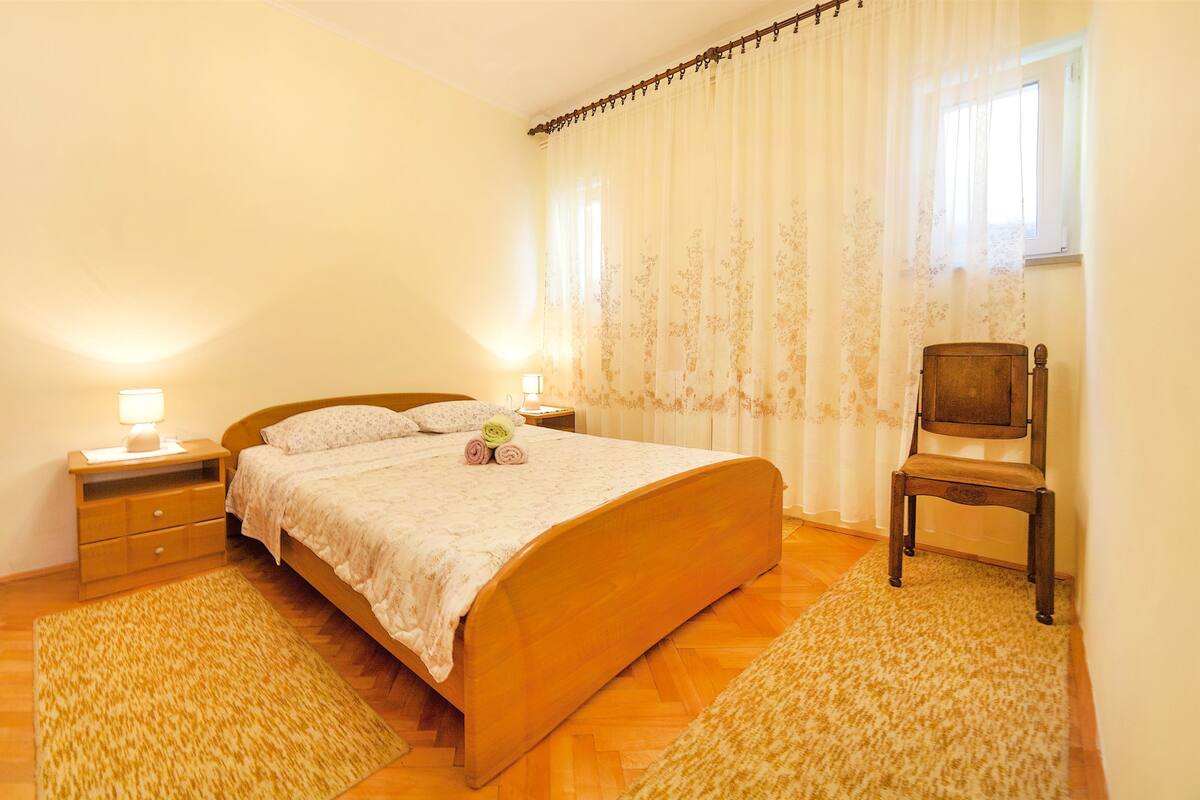 A bedroom with a wooden bed, two bedside tables with lamps, a chair, and a window with curtains.