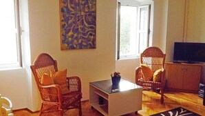 Un salon avec des fauteuils en osier, une table basse en verre, un téléviseur à écran plat et un tableau accroché au mur.