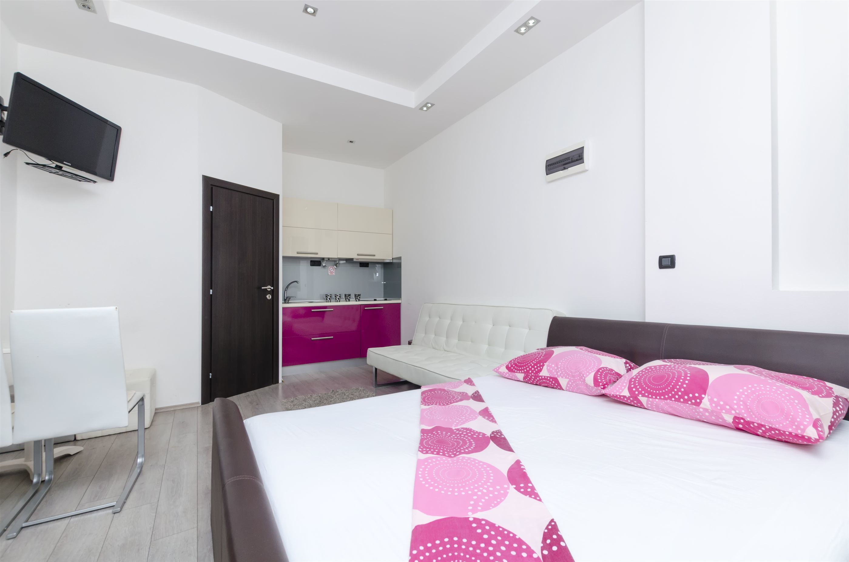 A modern hotel room with a large bed, a TV mounted on the wall, a small kitchenette, and a white chair.