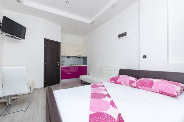 A modern hotel room with a large bed, a TV mounted on the wall, a small kitchenette, and a white chair.