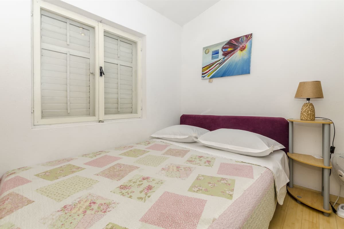 Una camera da letto con un letto, una finestra con persiane, un quadro appeso al muro e un comodino con una lampada.