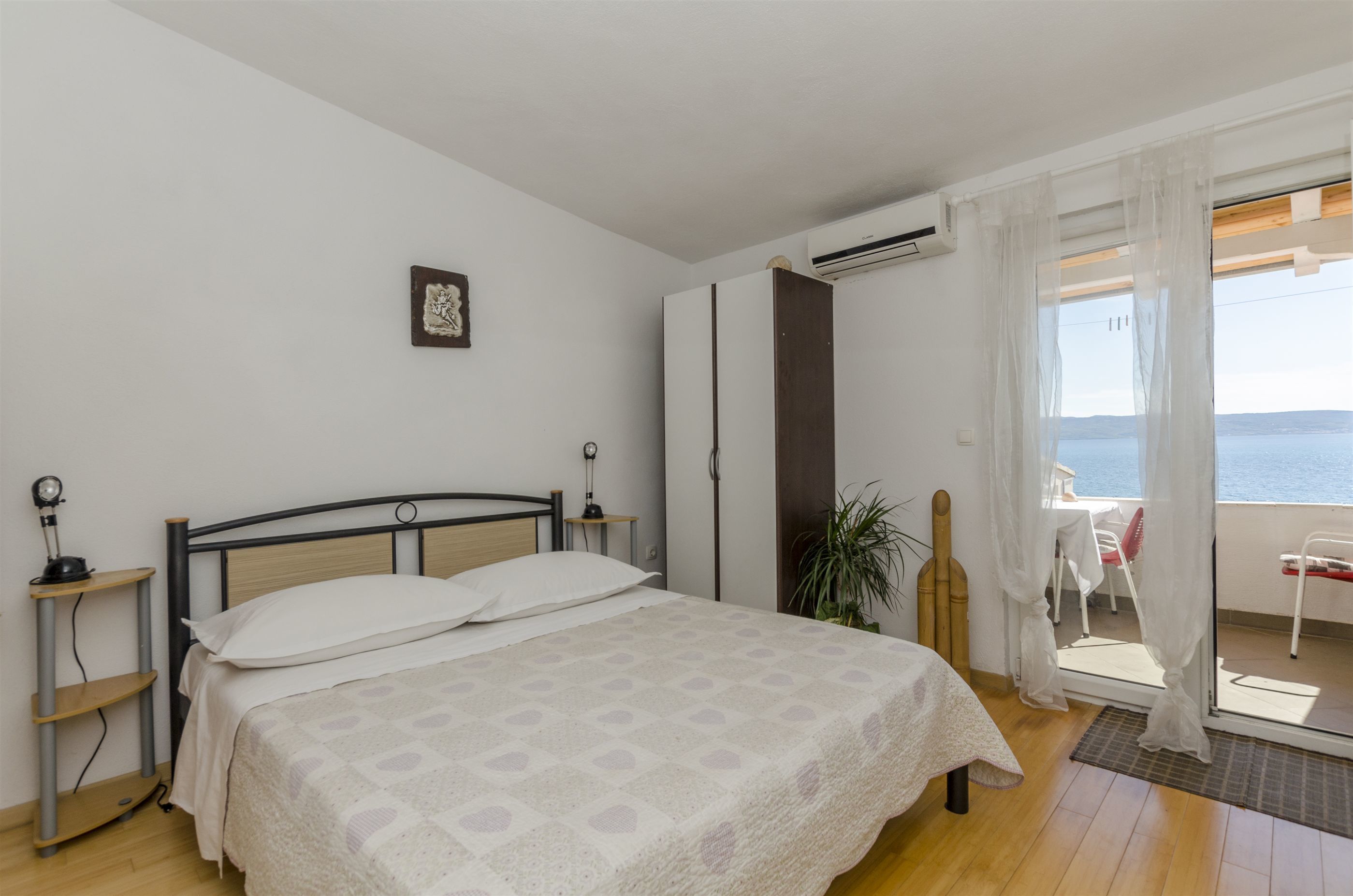 A bedroom with a bed, bedside tables, a potted plant, and a balcony with a view of the sea.