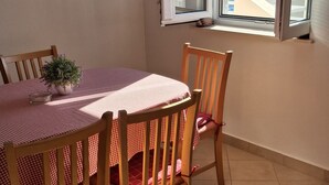 Ein Essbereich mit einem runden Tisch, Stühlen, einem Blumentopf und Blick auf ein Gewässer durch ein offenes Fenster.