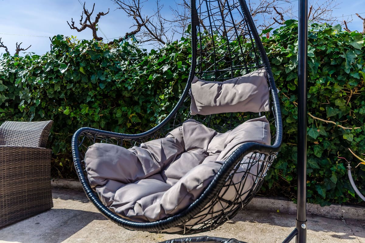 A hanging chair with cushions, a wicker table, and a hedge in the background.