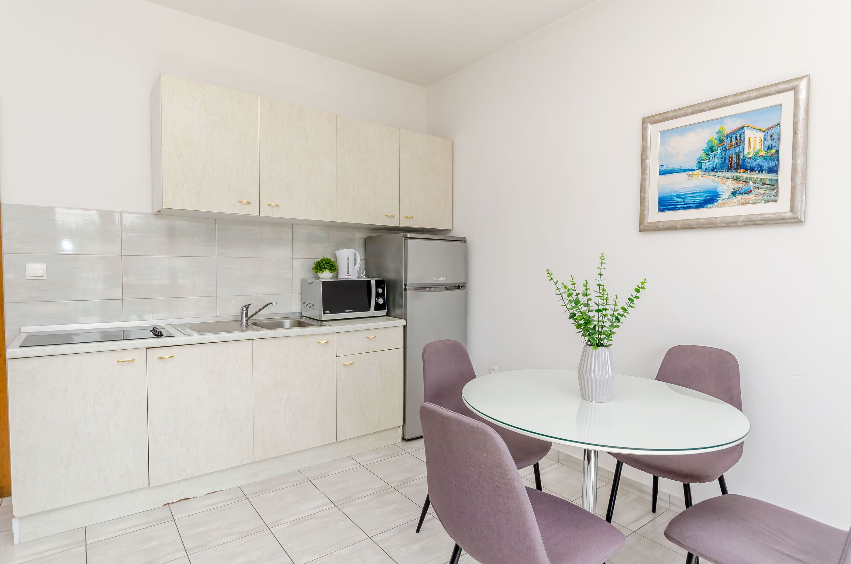 Eine moderne Küche mit weißen Schränken, einem Edelstahlkühlschrank, einer Mikrowelle und einem Spülbecken. Ein runder Tisch mit violetten Stühlen und eine Vase mit Grünpflanzen stehen dort. Ein gerahmtes Gemälde zeigt eine Küstenlandschaft, das an der Wand hängt.