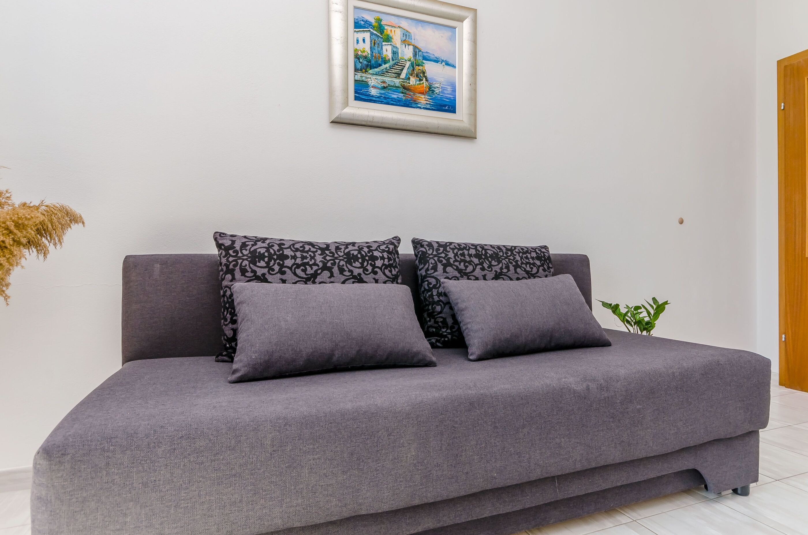 A grey sofa with patterned cushions, a framed painting of a coastal scene, and a small plant on the floor.