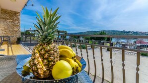 Un balcon avec une table sur laquelle se trouve un saladier rempli de fruits, notamment un ananas et des bananes, offrant une vue panoramique sur un paysage avec une étendue d’eau et des collines au loin.