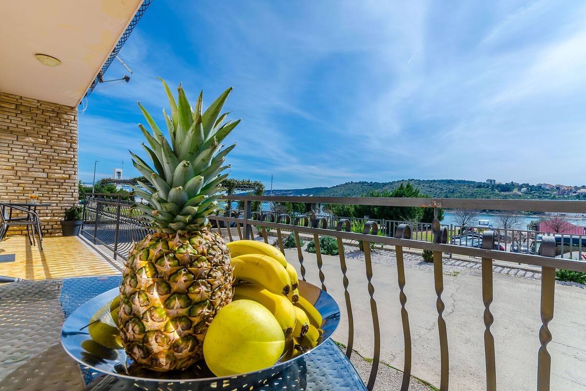 A balcony with a table holding a bowl of fruits, a brick wall, and a view of a distant landscape.
