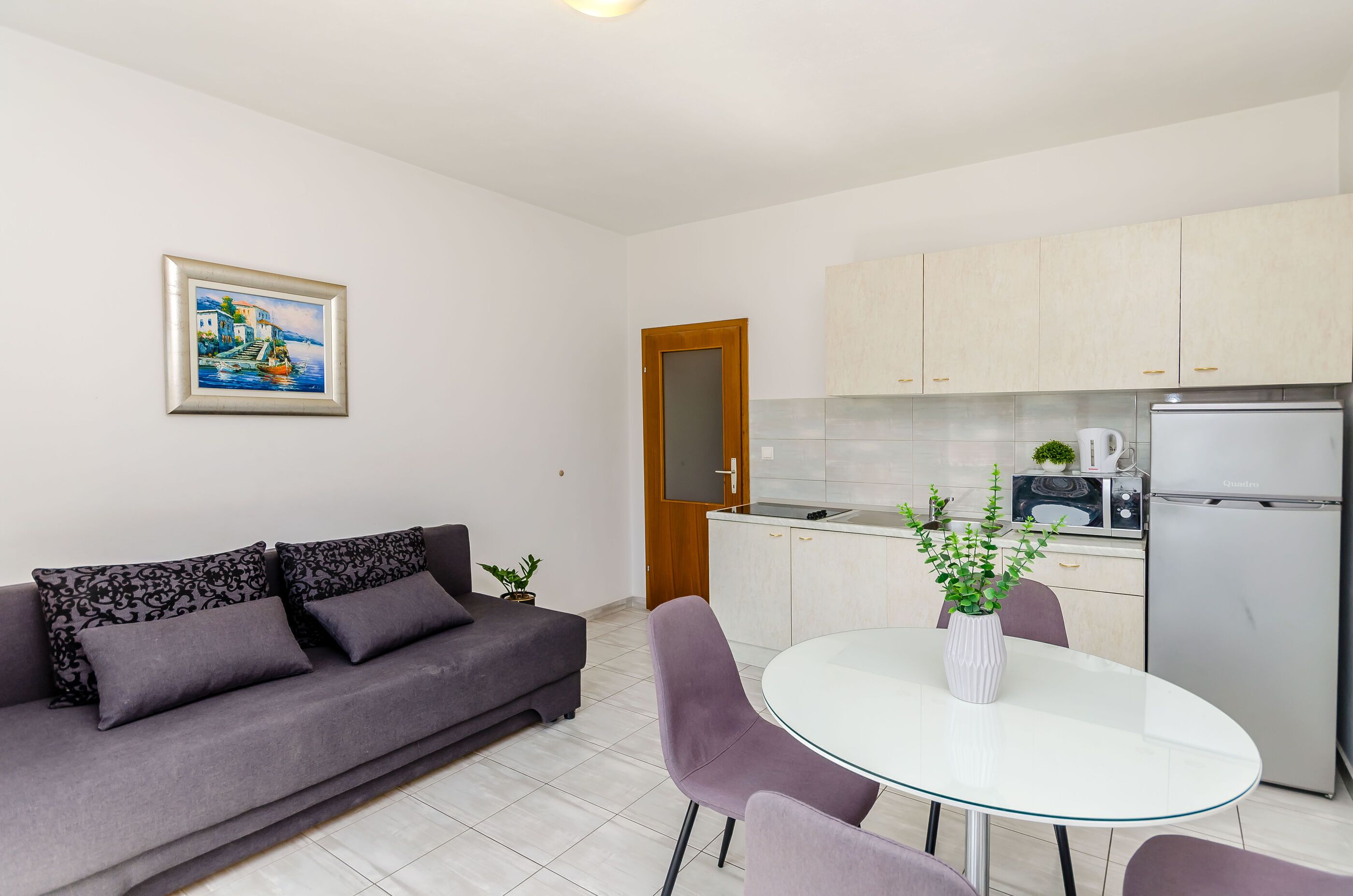 A modern living room with a sofa, a dining table with chairs, a kitchen area with cabinets and appliances, and a framed painting on the wall.
