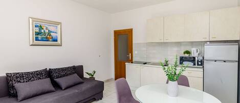A modern living room with a sofa, a dining table with chairs, a kitchen area with cabinets and appliances, and a framed painting on the wall.