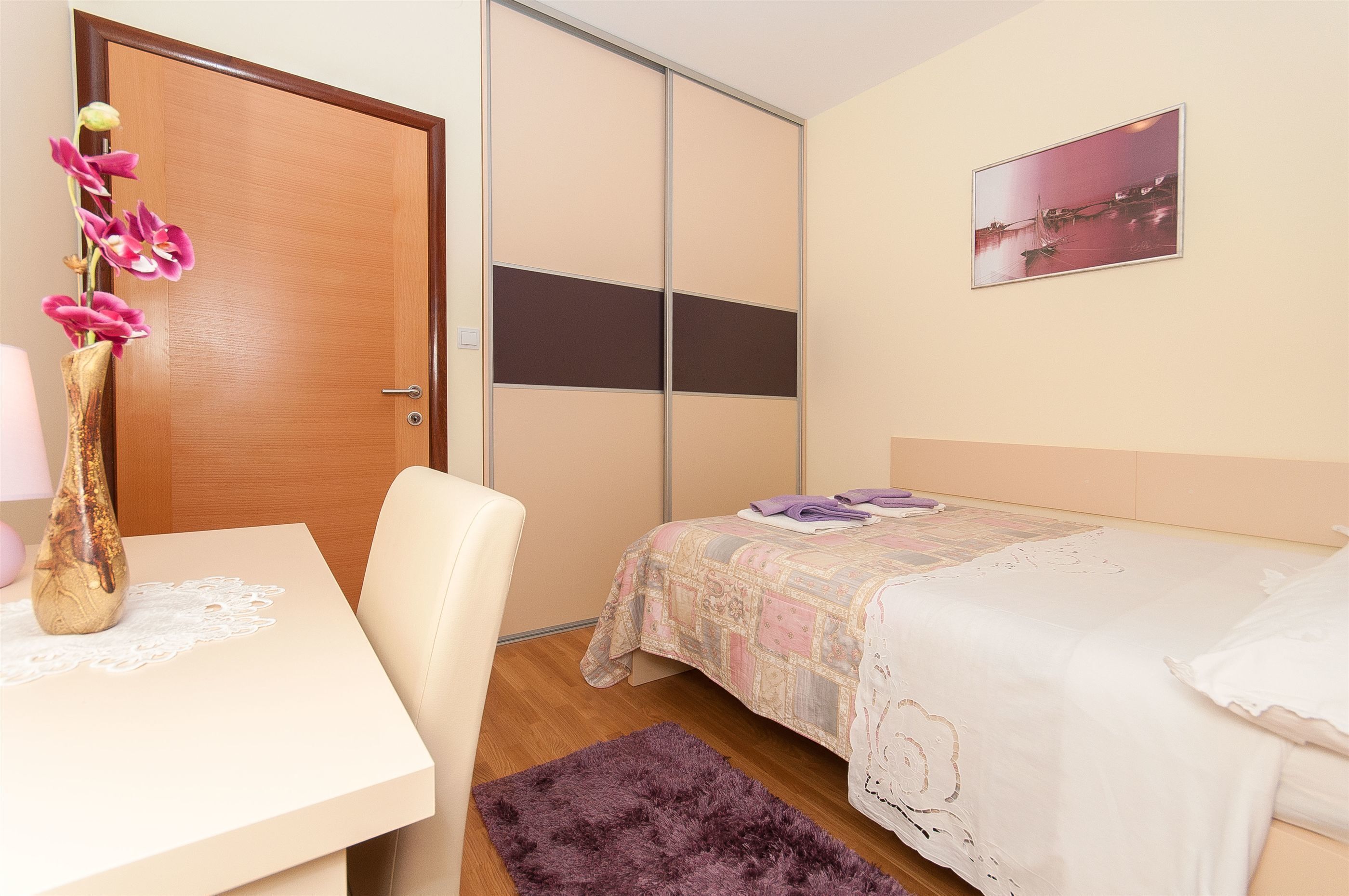 A bedroom with a bed, a desk, a chair, a door, and a painting on the wall.