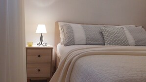 A neatly made bed with a white comforter and patterned pillows, a bedside lamp, and a wooden nightstand with drawers. - Apartments Mirjana (Milna)