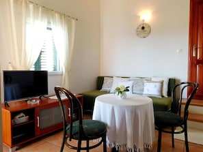 Una sala de estar con una televisión sobre un mueble de madera, un sofá verde con cojines estampados, una mesa redonda con mantel blanco y dos sillas.