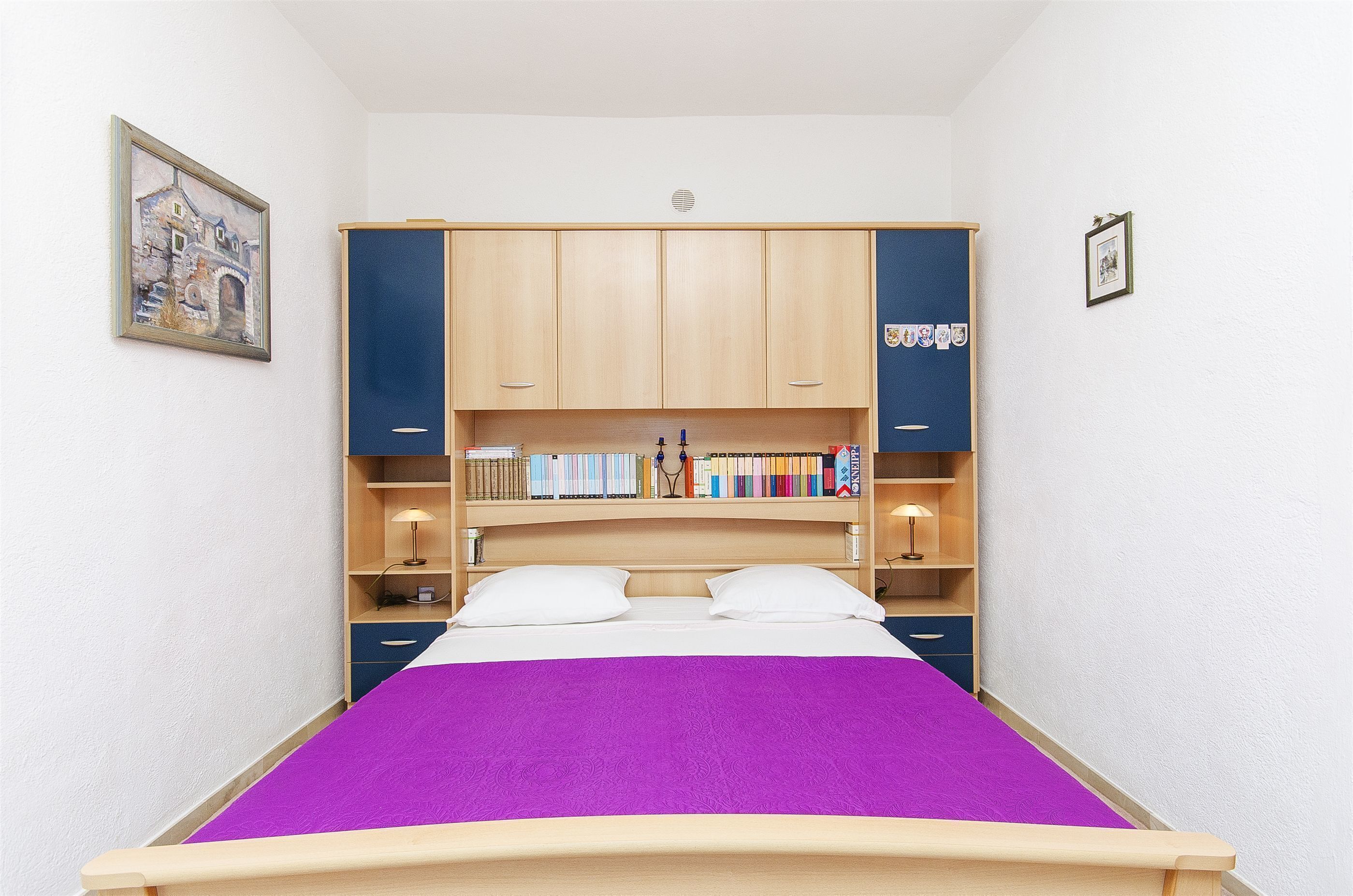 A bedroom with a bed, wooden wardrobe, shelves with books, and a framed picture on the wall.