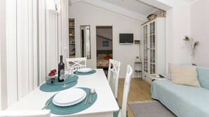 A dining area with a white table set for two, a television mounted on the wall, and a light blue sofa in the background. - Apartment Taida (Split)