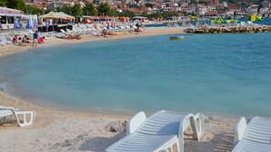 A beach with white lounge chairs, a clear blue sea, and a town in the background. - Apartment Marin (Trogir)