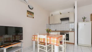 A compact kitchen with white cabinets, a stove, and a refrigerator. There is a dining table with a checkered tablecloth and chairs. A flat-screen TV is on a stand in front of the table. - Apartments Gloria (Rogoznica)
