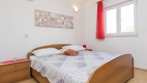 A bedroom with a wooden bed, a nightstand, a wall-mounted light, a window with a red curtain, and a floral artwork on the wall. - Apartments Gloria (Rogoznica)
