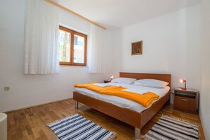 A bedroom with a wooden bed, white bedding, a window with curtains, a bedside table with a lamp, and a painting on the wall. - Apartment Ante Tonci (Makarska)