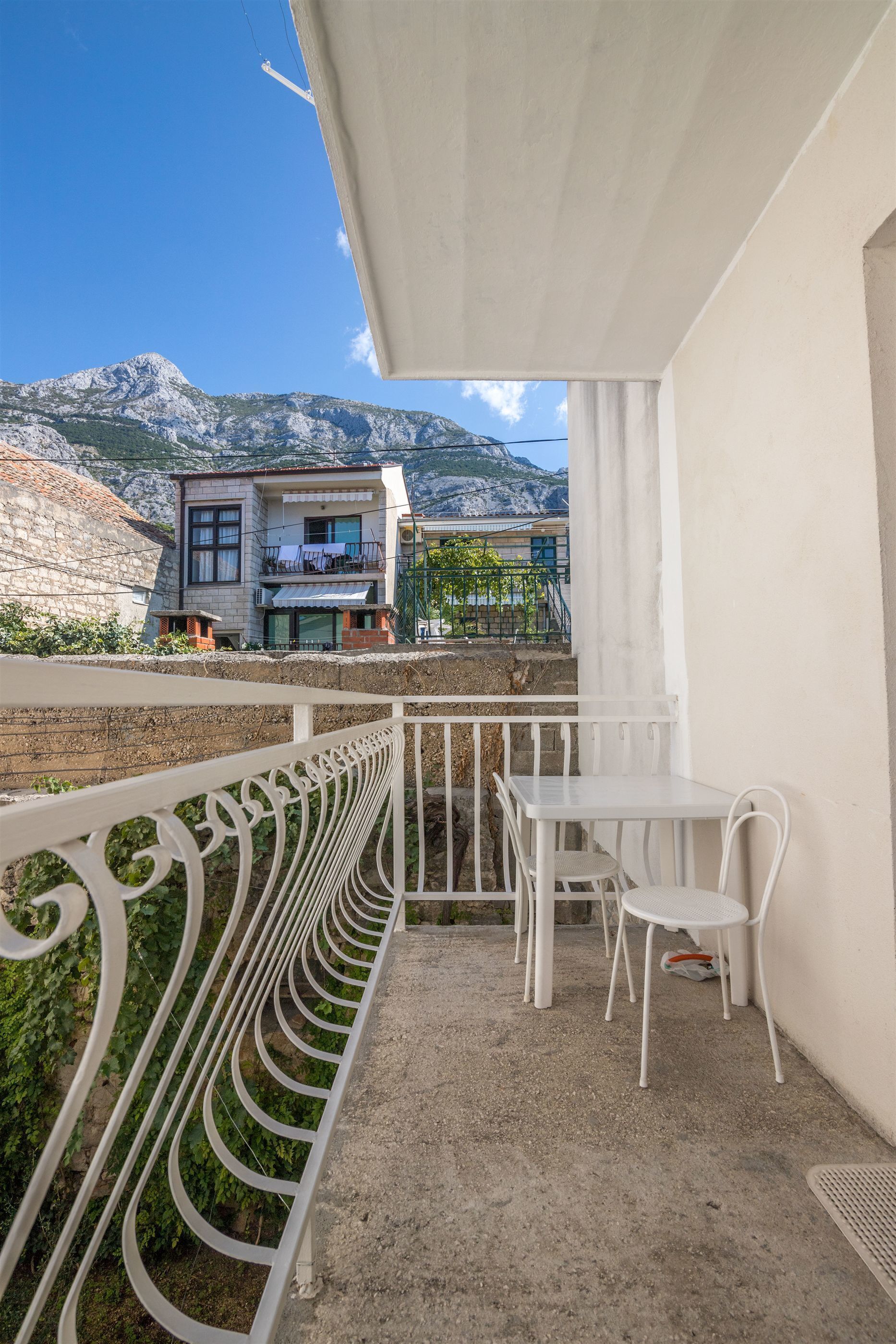 A balcony with a white metal railing, a white table, and two white chairs. In the background, there is a building with a balcony and a mountain.