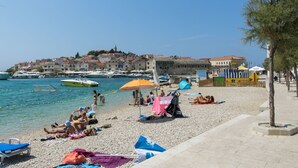 Una playa con gente tomando el sol y nadando, barcos en el agua y edificios al fondo.