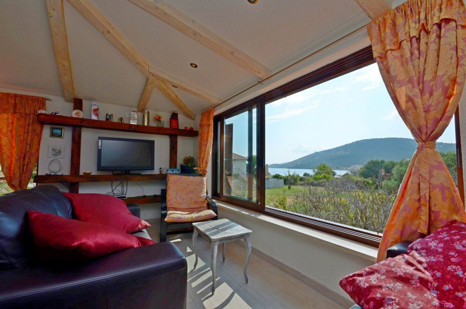A living room with a TV, a sofa, a chair, and a view of a body of water and hills.