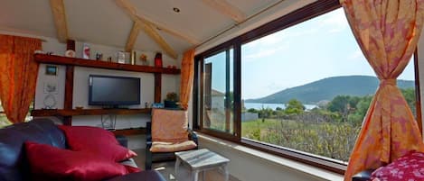 A living room with a TV, a sofa, a chair, and a view of a body of water and hills.