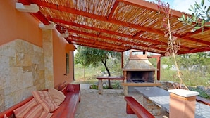 Une terrasse extérieure dotée d’un sol en pierre, de bancs en bois, d’une cheminée en pierre et d’une structure à toit de chaume.