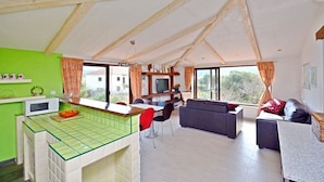 Un loft spacieux comprenant une cuisine, un coin repas et un salon. Il se distingue par son plafond haut avec des poutres apparentes, de grandes fenêtres et une vue sur l’extérieur.