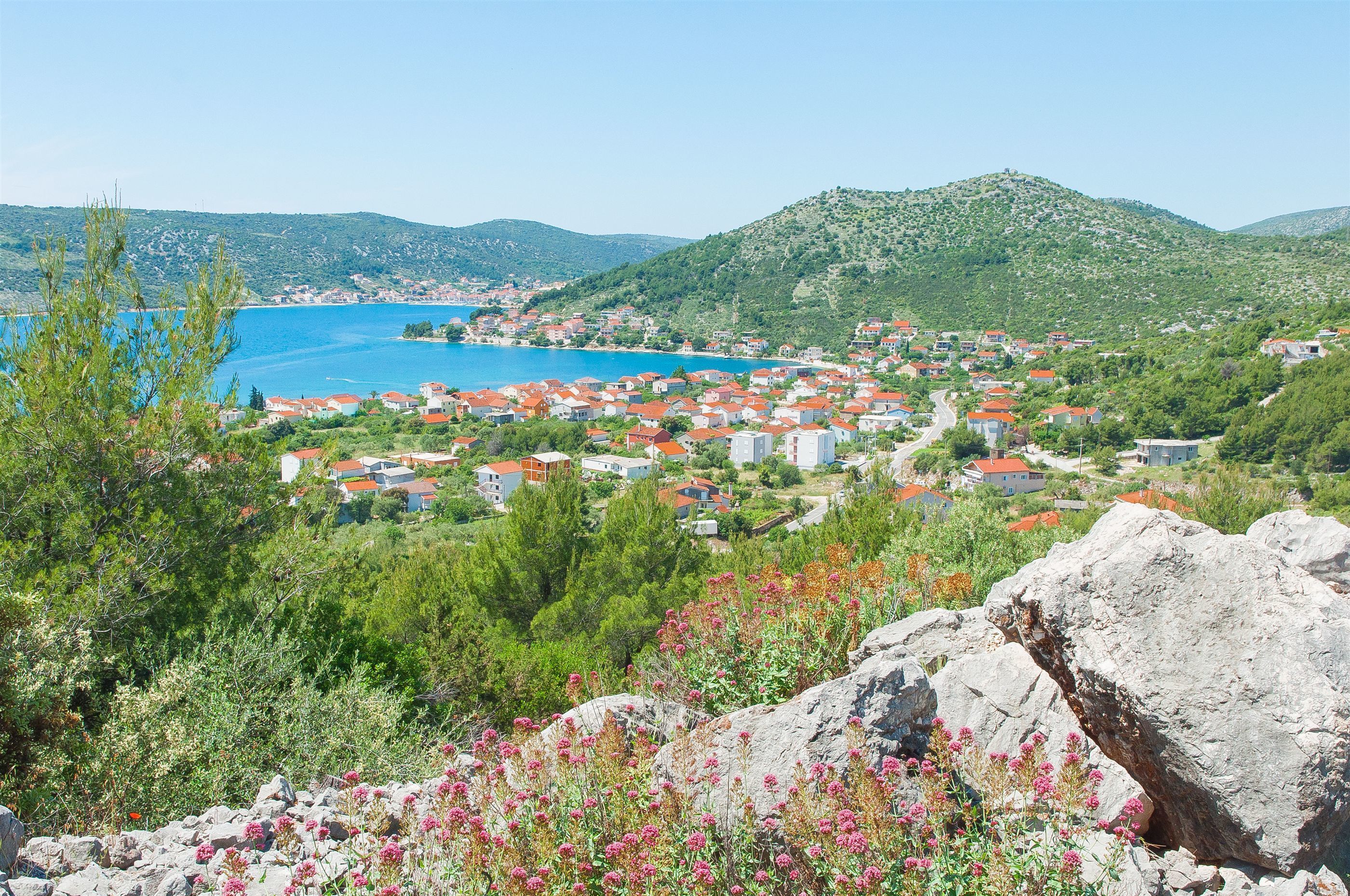 Ein Küstdorf mit Häusern, blauem Meer und grünen Hügeln im Hintergrund.