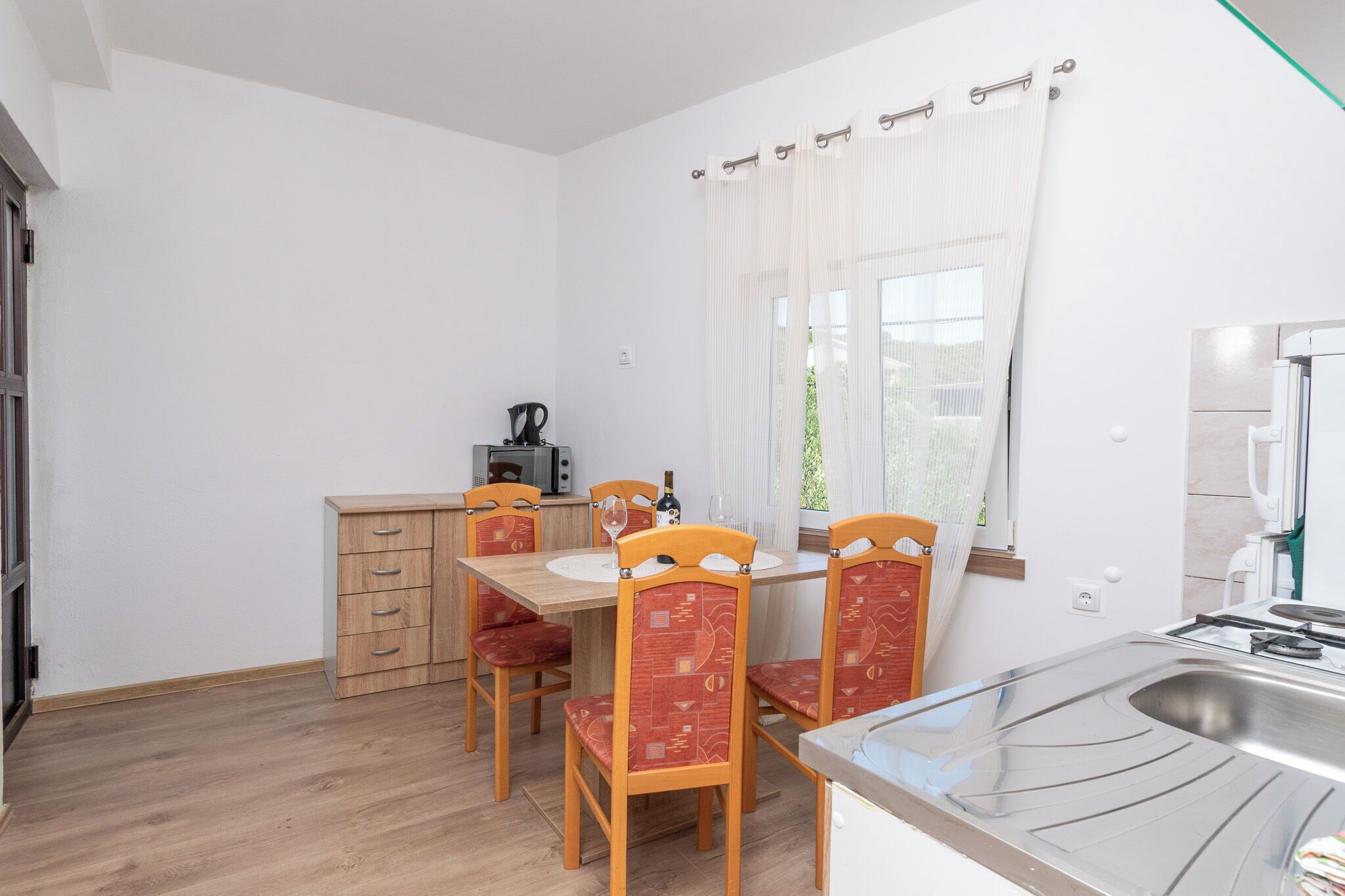 A compact kitchen with a dining table, chairs, a small cabinet, a window with curtains, and a sink.