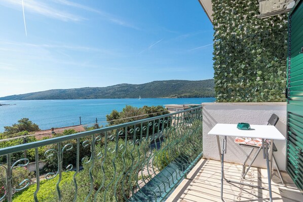 A balcony with a view of a lake and mountains, a table with a chair, and a green wall with foliage.