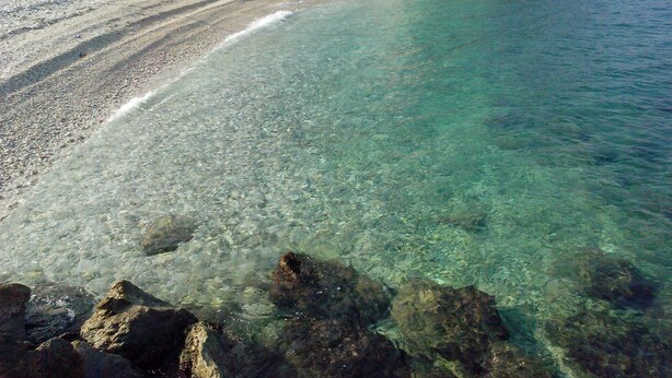 A beach with clear water, pebbles, and rocks. There is a building with windows in the background. - Apartments Anita (Slatine)