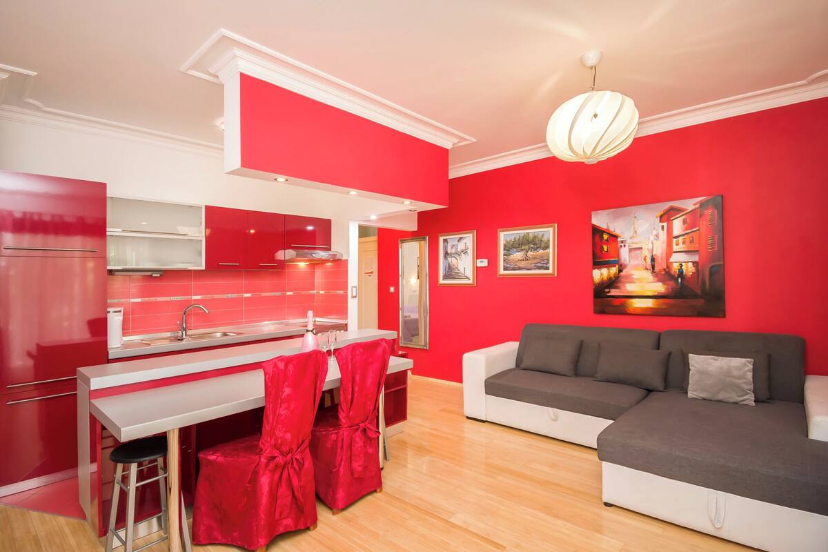 A modern kitchen with red cabinets, a white countertop, and a bar area. There is a gray sofa and a painting on the wall.