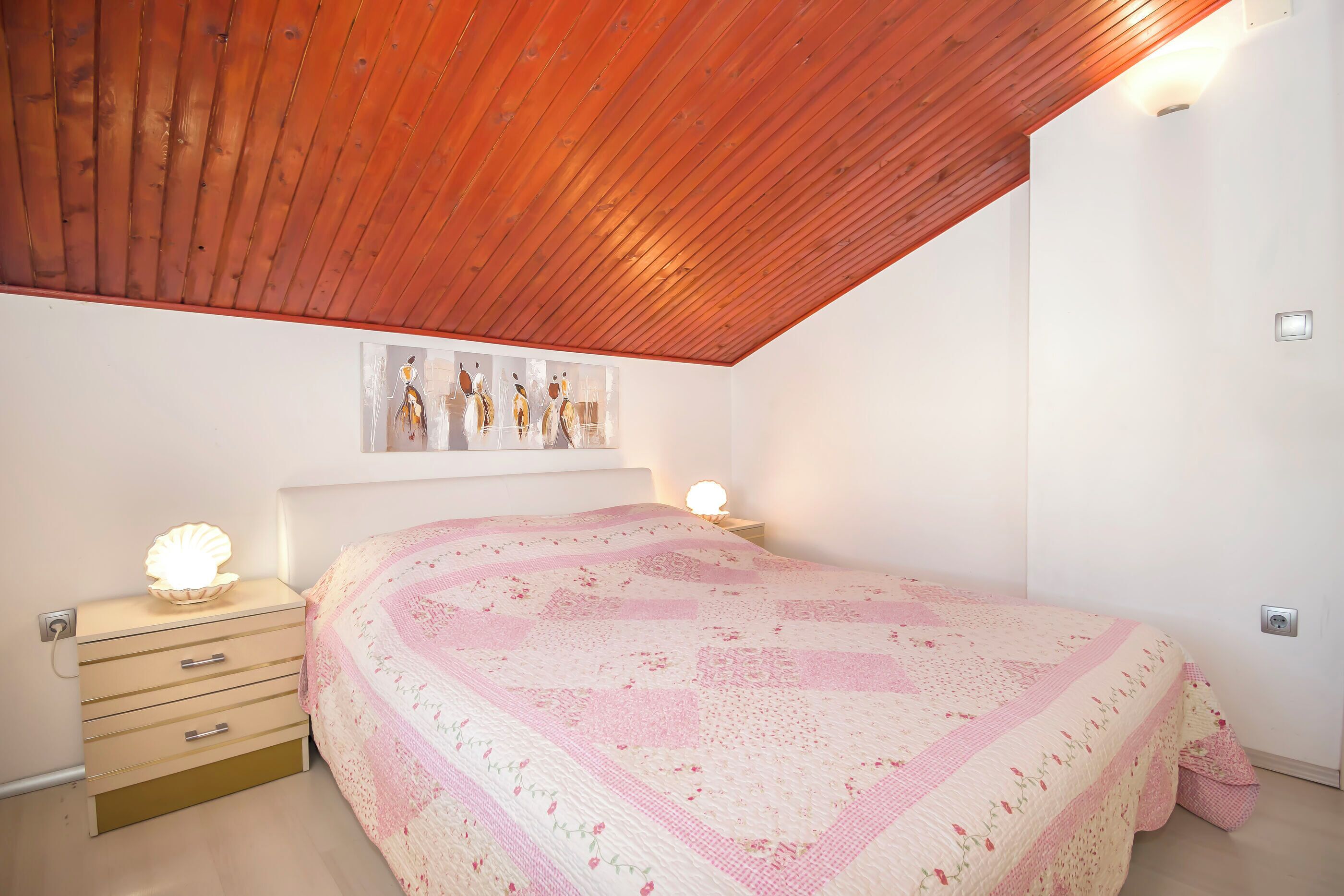 Un dormitorio con techo de madera, una cama con colcha rosa, l ámparas de noche y un accesorio de luz empotrado en la pared.