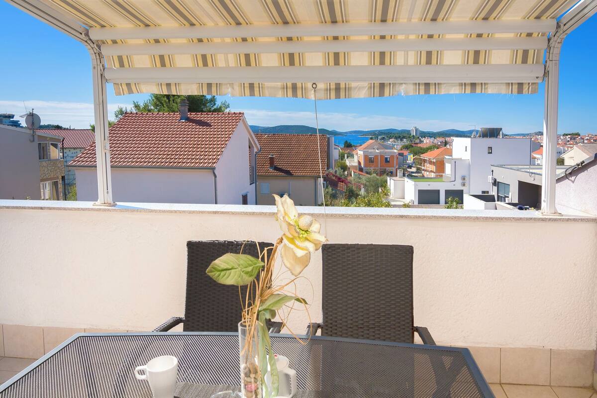 A covered outdoor seating area with a table and chairs, a vase with flowers, and a view of buildings and hills in the background.