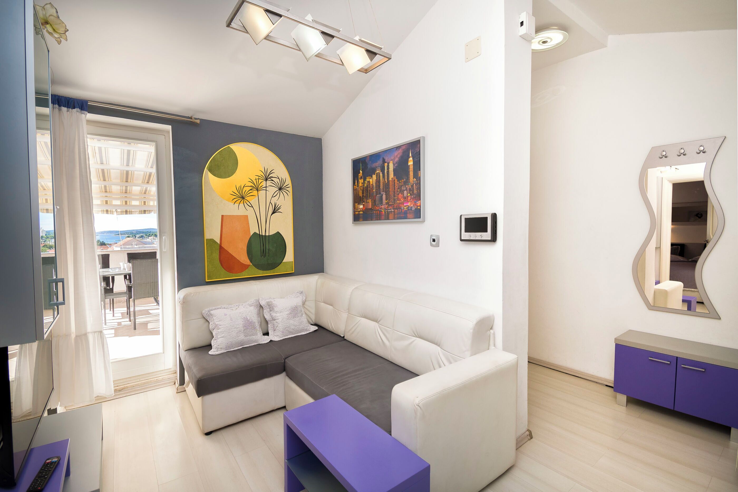 Una sala moderna con un sofá blanco, una mesita auxiliar morada y un cuadro de un paisaje urbano.