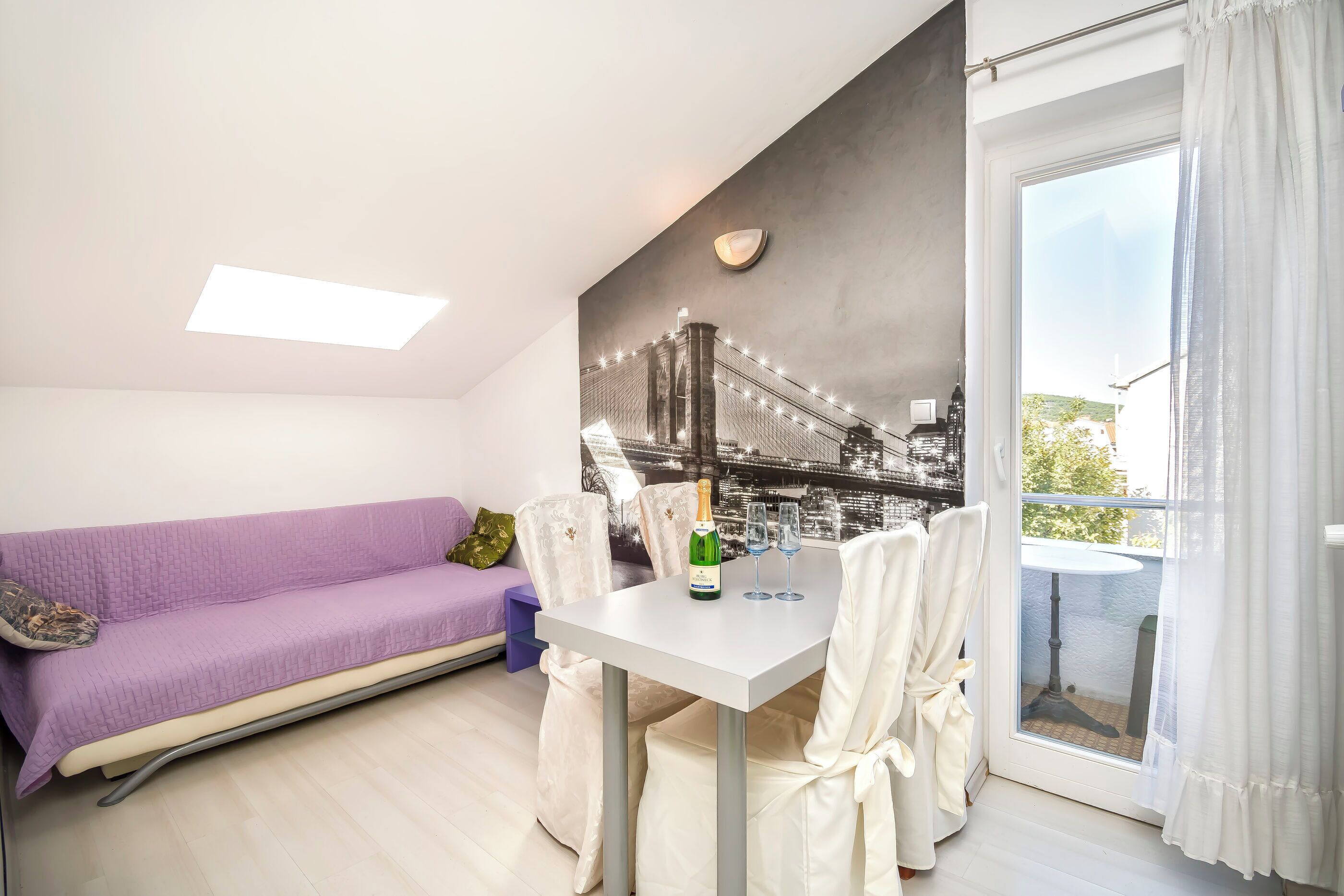 Et moderne værelse med en lilla sofa, et hvidt bord med en flaske og glas, samt et stort vindue med gardiner.