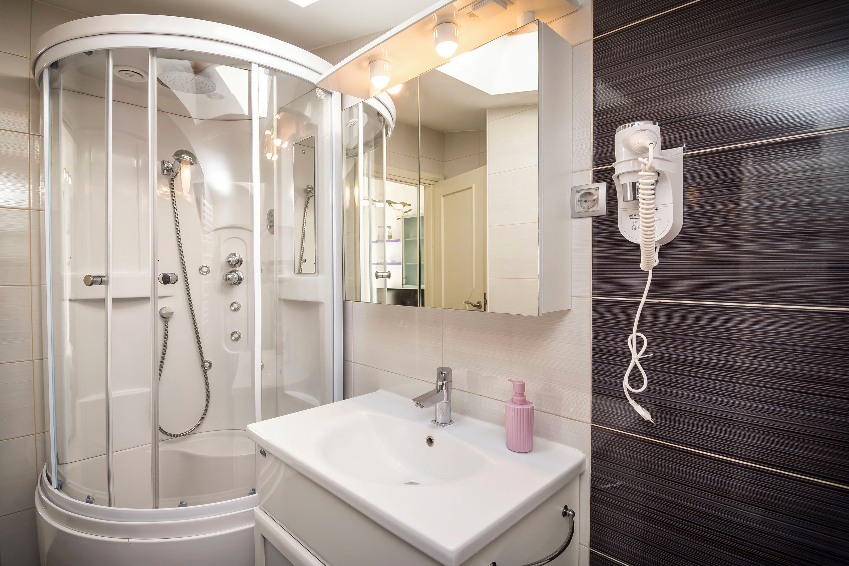 A modern bathroom with a glass-enclosed shower, a white sink, a mirror, and a hairdryer mounted on the wall.