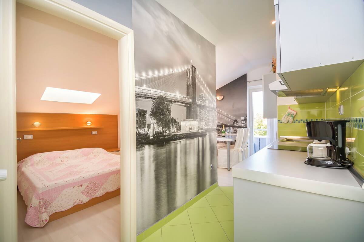 A compact room with a bed, a kitchenette, and a large window with a cityscape view.