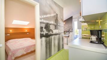 A compact room with a bed, a kitchenette, and a large window with a cityscape view.