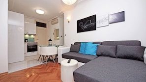 Una sala moderna con un sofá gris, una mesita redonda y una silla blanca. En el fondo, se ve una cocina con armarios blancos y una estufa.