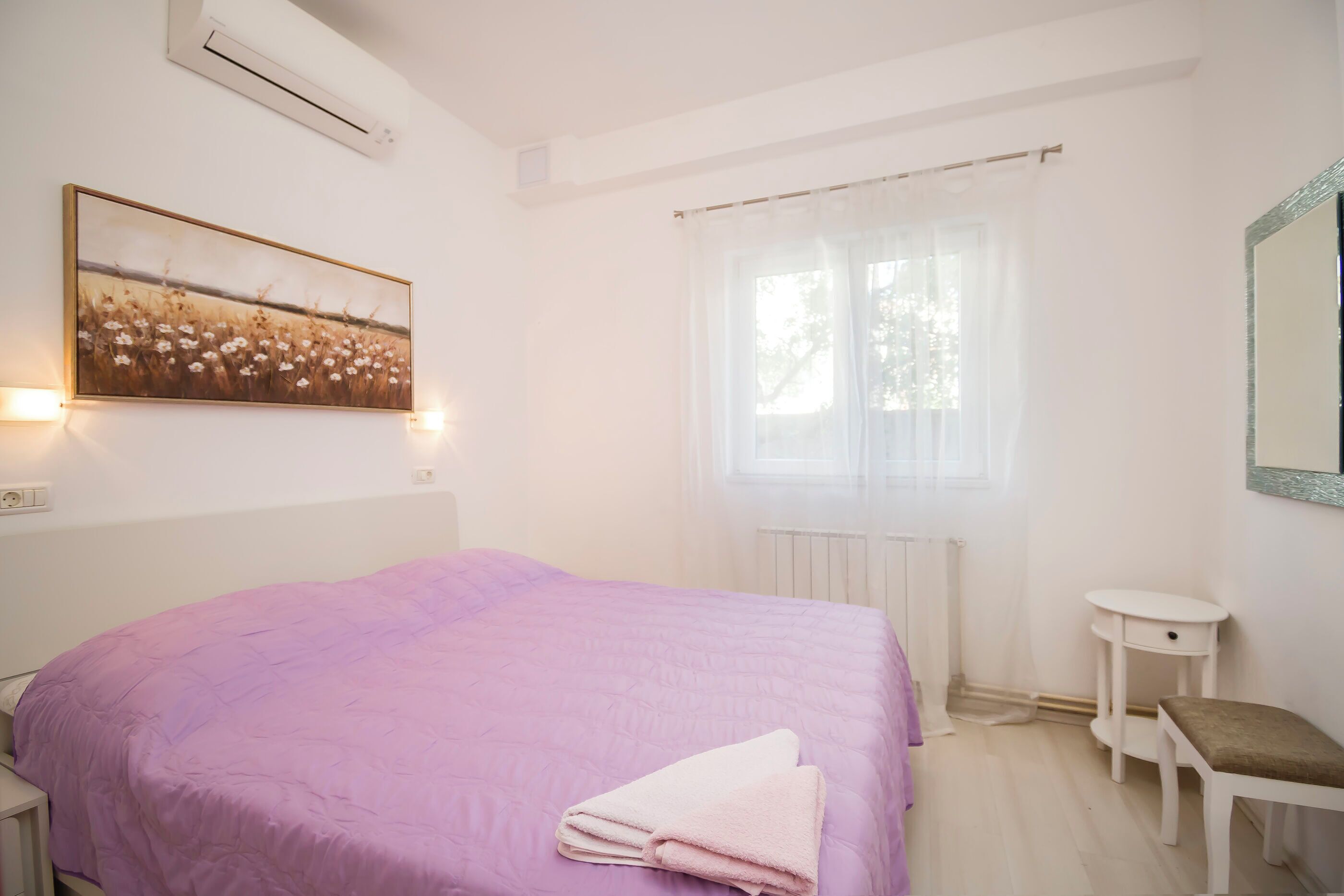 Un dormitorio con cama, mesita de noche, una pequeña mesa, ventana con cortinas y aire acondicionado instalado en la pared.