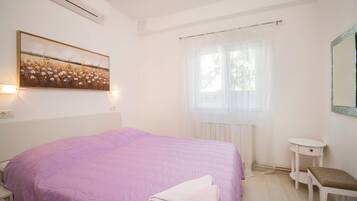 Un dormitorio con cama, mesita de noche, una pequeña mesa, ventana con cortinas y aire acondicionado instalado en la pared.