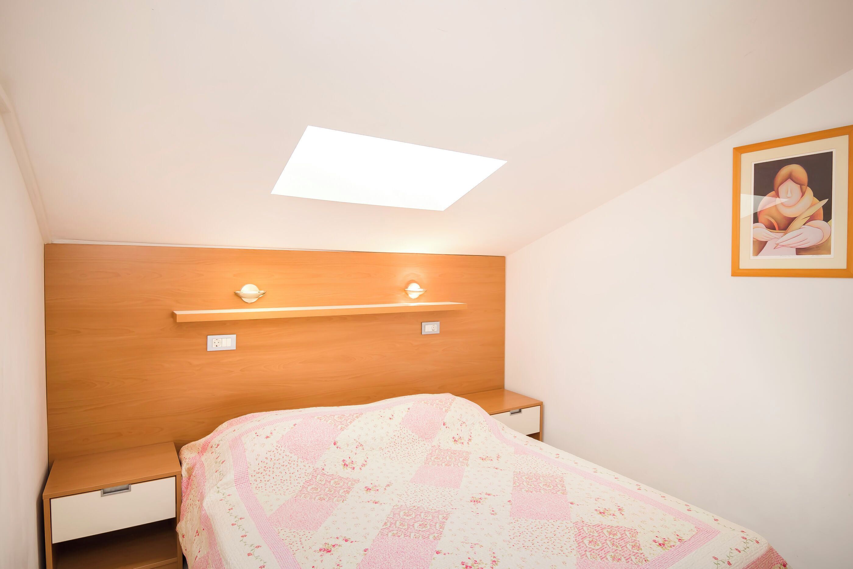 A bedroom with a wooden headboard, a bed with a patterned quilt, a bedside table, a nightstand, a small shelf, and a framed picture on the wall.
