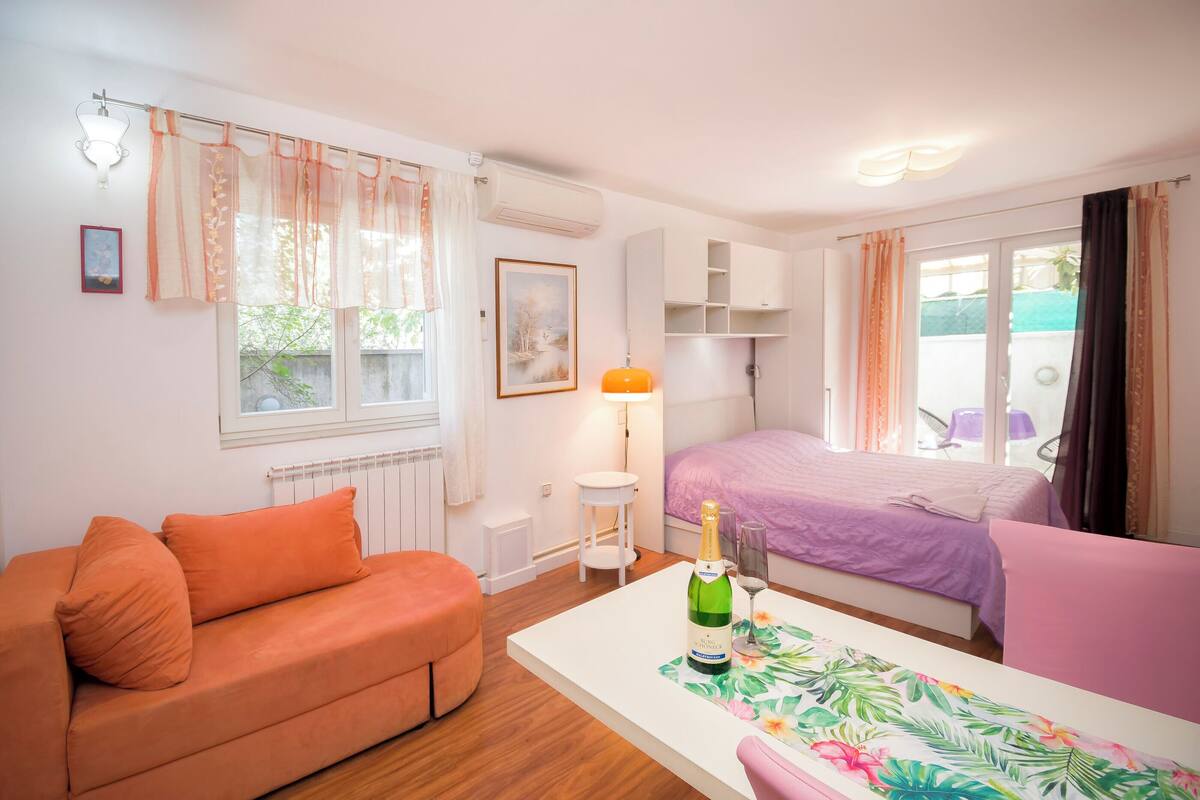 A room with a sofa, a bed, a table with a bottle and glass, and a window with curtains.