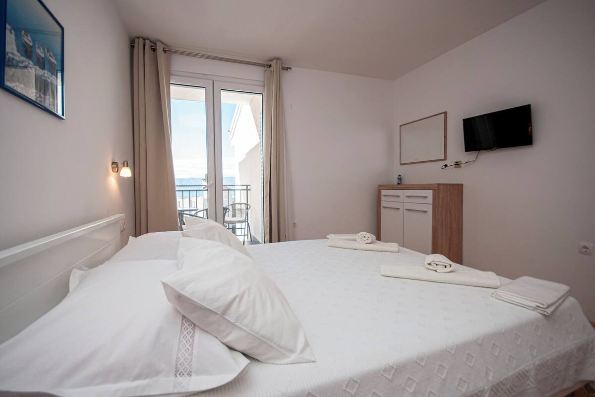 Een hotelkamer met een bed, een aan de muur bevestigde televisie, een klein bureau met een spiegel, en een balkon met een stoel.
